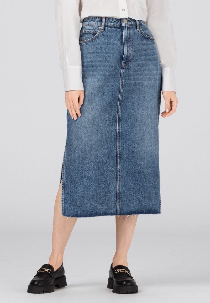 Kvinna iklädd en midilång blå jeanskjol med sidofrånskärning, vit knäppt skjorta och svarta loafers med detaljer i guldkedja.