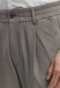 Gros plan de la main d'une personne reposant sur un pantalon gris à motif chevrons taille haute, avec un bouton et une boucle de ceinture, portant une chemise blanche.