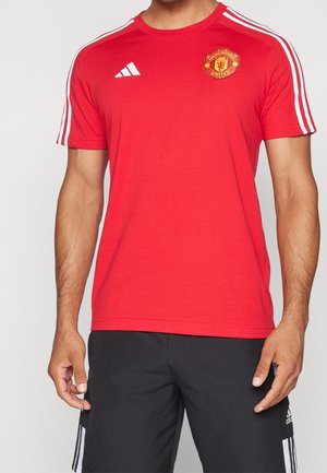 Homme portant un maillot de sport rouge à manches courtes de Manchester United avec un logo Adidas blanc et des épaules rayées, porté avec un short noir Adidas.