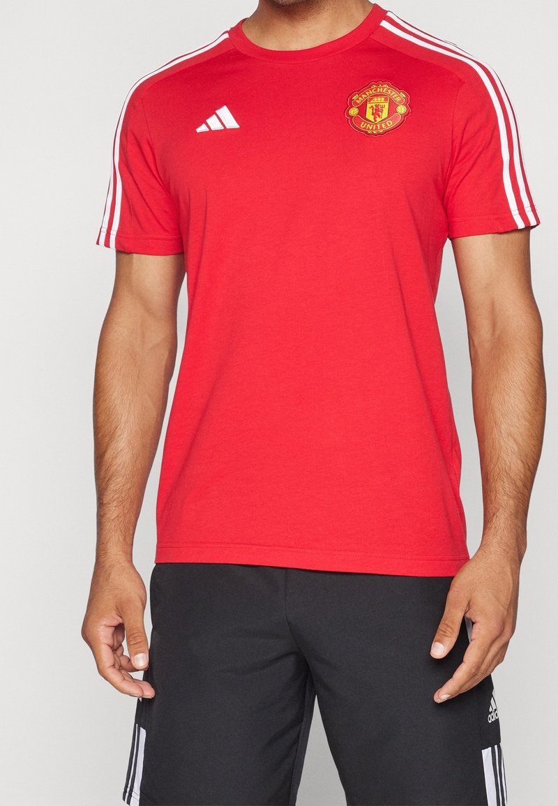 Homme portant un maillot de sport rouge à manches courtes de Manchester United avec un logo Adidas blanc et des épaules rayées, porté avec un short noir Adidas.