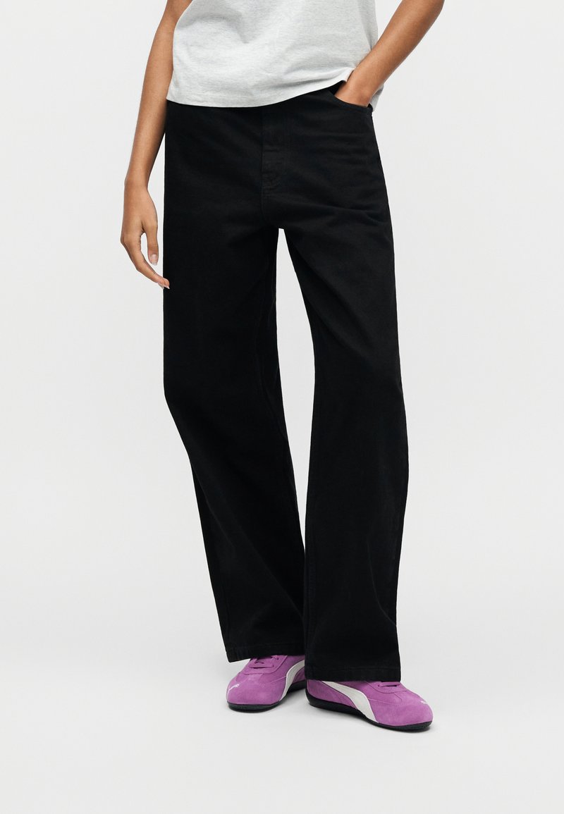 Zwarte broek met wijde pijpen van een gladde stof, voorzien van steekzakken aan de voorkant en een klassieke tailleband, gecombineerd met paarse sneakers.