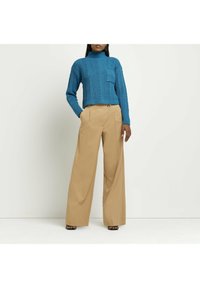 Maglione turtleneck in teal lavorato a maglia con una tasca frontale, abbinato a pantaloni beige a gamba larga con bottone frontale e pieghe.