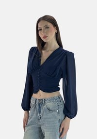 Himmelblaues, verkürztes Blusenoberteil mit langen Puffärmeln, gerafften Details und vorderem Knopfverschluss, kombiniert mit hochgeschnittenen, hellen Jeans.