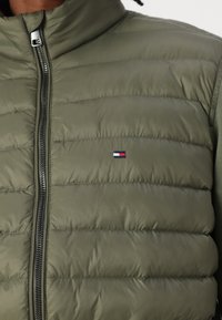Doudoune verte olive avec un col montant, présentant des coutures horizontales, une fermeture éclair complète et un petit patch logo sur la poitrine.