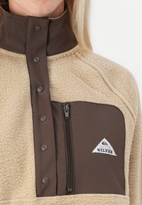 Fleece jacket met een beige lichaam en een bruine drukknoopkraag, voorzien van een ritsborstzak met een driehoekig logo en gestructureerde stof.