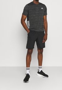 Musta urheilupaita, jossa on teksturoitu kangas, valkoinen Adidas-logo, yhdistetty mustiin shortseihin ja mustiin urheilukenkiin, joissa on valkoiset raidalliset sukat.