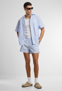 Chemise à rayures bleu clair à manches courtes, débardeur blanc, shorts assortis et sabots beiges ; modèle portant des lunettes de soleil sombres et des chaussettes blanches.