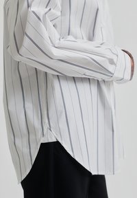 White striped shirt with vertical navy stripes, featuring long sleeves, a curved hem, and soft cotton fabric for a relaxed fit.