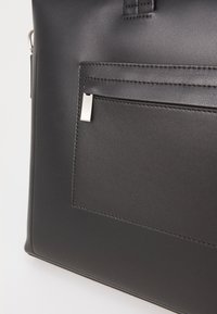 Čierna kožená taška na rameno so prednou vreckom, ktoré má strieborný kovový zips a kontrastné stehy, čo zdôrazňuje štýlový, moderný dizajn.