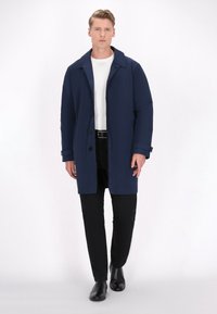 Mørkeblå trenchcoat med klippet krave, knaplukning og sidelommer, parret med en hvid t-shirt og sorte bukser, båret med sorte sko.