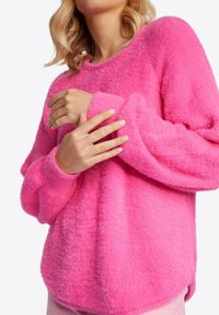 Maglione rosa fuzzy con una vestibilità ampia, scollo rotondo e maniche lunghe. La texture appare morbida e leggermente pelosa.