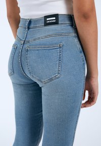 Jeans de mezclilla de tiro alto en azul claro con un diseño ajustado, con una etiqueta negra en la parte trasera y dos bolsillos traseros. Textura suave.