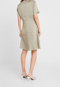 Ärmelloses Kleid in Creme mit geometrischem Muster in Gold und Schwarz, knielang, fließende Silhouette, kombiniert mit weißen Riemchenschuhen.