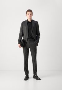 Grijs pak gemaakt van gestructureerde stof, gecombineerd met een zwart shirt en zwarte schoenen. Een zwarte clutch wordt vastgehouden in de linkerhand; een moderne, op maat gemaakte pasvorm.