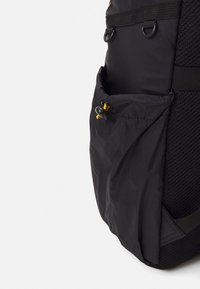 Svart ryggsekk med et teksturert mesh-panel, med en sidelomme som er sikret med en snorlukking. Holdbar stoffkonstruksjon.
