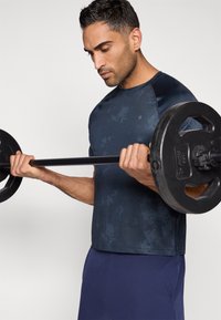 Sötétkék sportpóló texturált mintával, miközben egy kerek fekete súlyt emel. A pólón lekerekített gallér és rövid ujjak találhatók.