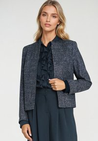 Blondynka w teksturowanej szarej kurtce nosząca czarną bluzkę z falbanami i pasujące czarne spodnie z wysokim stanem na jednolitym tle.