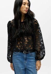 Sort gennemsigtig bluse med indviklet blomster- og blondenetmønster, puffede ærmer og knappedetaljer. Kombineret med blå denimjeans og sort bælte.