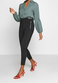 Grön texturerad blus med knäppning och samlade ärmar, parat med svarta figurnära byxor med zipfickor och röda remsandaler.