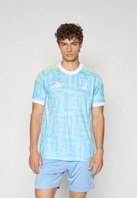 Camiseta de fútbol de manga corta de color azul claro con un patrón en espiral, detalles blancos y acentos amarillos en los hombros; combinada con pantalones cortos de color azul claro.