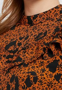 Blusa arancione con motivi floreali e animalier in nero. Tessuto testurizzato con dettagli a volant lungo il colletto e le spalle.