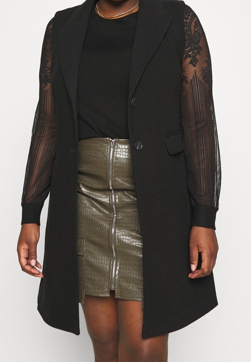 Blazer noir sans manches porté sur un top transparent à manches longues, jupe en simili cuir vert olive avec fermeture éclair frontale, sur une personne devant un fond blanc.