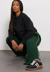 Svart knapp-up sweatshirt, gröna vida byxor med vita sidostripes och svarta sneakers med vita detaljer. Slät tyg och avslappnad passform.