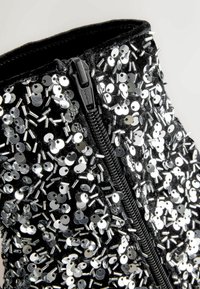 Mustmustest musta, mis on kaetud metalliliste hõbedaste litrite ja helmedega, millel on külgvetti. Pind on tekstuurne ja peegeldav.