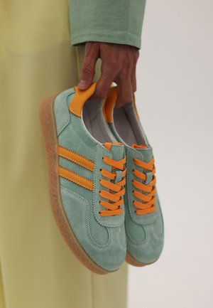 Hånd, der holder lyse turkis sneakers med orange snørebånd og striber mod en neutral baggrund.