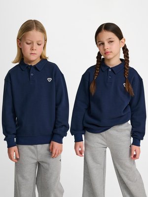 Zwei Kinder stehen nebeneinander, sie tragen marineblaue Poloshirts mit langen Ärmeln und graue Jogginghosen vor einem schlichten weißen Hintergrund.