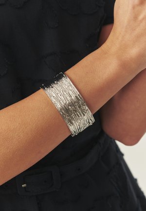 Široka teksturirana srebrna zapestnica, nošena na zapestju ob črnem teksturiranem topu in pasu.
