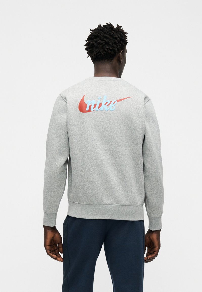 Man with short curly hair wearing a grey sweatshirt with a large red and blue Nike logo on the back, paired with dark pants.