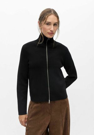 Femme aux cheveux châtain clair portant un pull noir zippé et un pantalon marron, debout devant un fond blanc uni.