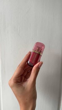 Una mano che tiene una piccola bottiglia cosmetica rossa etichettata "SuperDewy Blush Burst" con un tappo rosa traslucido su uno sfondo semplice.