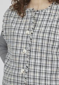 Rutet stofvest i gr å, sort og hvid med en frayed tekstur. Har runde knapper og en rund halsudskæring, hvilket giver et lagdelt look.