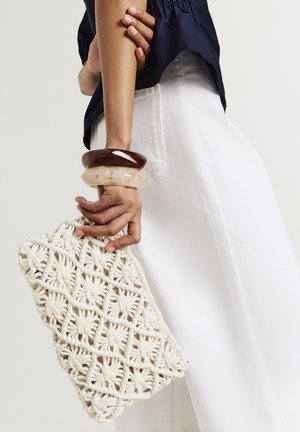Borsa a mano in macramè color crema con intricati motivi floreali, tenuta in una mano adornata da tre bracciali in resina sovrapposti.