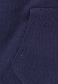 Navyblauwe katoenstof met een gestructureerd oppervlak, voorzien van een gebogen zak omlijnd door een subtiele naad. Geen prominente patronen of accenten.