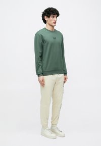 Zöld hosszú ujjú, kerek nyakú pulóver, krémszínű melegítőnadrággal, melyen fekete oldalsó nyomat található, fehér sportcipővel kiegészítve.