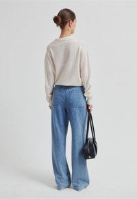 Světlý béžový svetr s otevřeným pleteným vzorem, doplněný volnými modrými džínami. Modelka drží malou černou kabelku se dvěma popruhy.