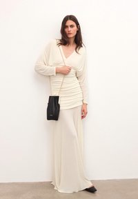 Kreminės spalvos ilga suknelė su sulenkta medžiaga, V formos iškirpte ir ilgas rankovėmis. Modelis laiko juodą rankinę su grandinėlės diržu, turinčia lygų paviršių.