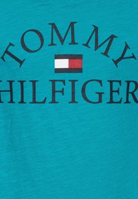 Tyrkysové bavlněné tričko s velkým černým textem "TOMMY HILFIGER" a malou vlajkovou logem v červené, bílé a modré barvě umístěnou pod ním.