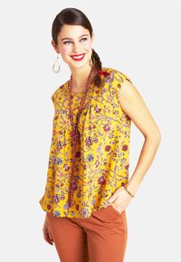 Gelbe Bluse mit Blumenmuster und kurzen Ärmeln, verziert mit lila und roten Motiven. Kombiniert mit brauner Hose und Perlenaccessoires.