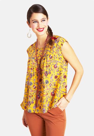 Gelbe Bluse mit Blumenmuster und kurzen Ärmeln, verziert mit lila und roten Motiven. Kombiniert mit brauner Hose und Perlenaccessoires.