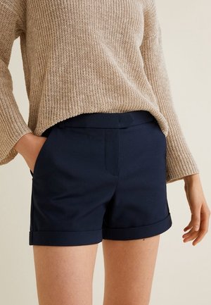 Pantaloni corti blu navy con un design su misura, caratterizzati da una texture liscia e orlo risvoltato. Abbinati a un maglione lavorato a maglia beige, morbido e largo.