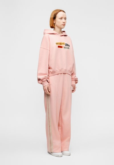 Ροζ φαρδιά μπλούζα με κουκούλα και γραφικό κείμενο, συνδυασμένη με αντίστοιχο παντελόνι φόρμας με φαρδιά μπατζάκια και ρίγες στο πλάι. Μαλακό ύφασμα και άνετη εφαρμογή.