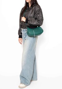 Borsa a tracolla verde in pelle con forma arrotondata e fondo piatto, dotata di una cerniera superiore e una finitura leggermente testurizzata. Indossata con un bomber jacket nero.