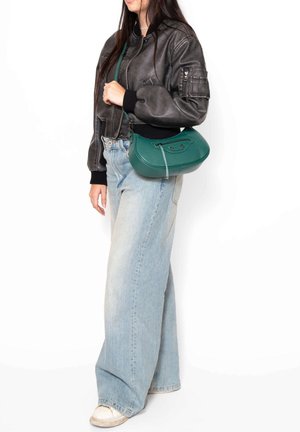 Grüne Leder-Crossbody-Tasche mit abgerundeter Form und flachem Boden, ausgestattet mit einem oberen Reißverschluss und einer dezenten strukturierten Oberfläche. Getragen mit einer schwarzen Bomberjacke.