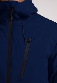 Marineblå jakke lavet af et glat, let materiale; har en høj krave, lynlåslommer med sorte metaldele og et minimalt design.