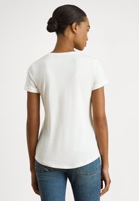 Sieviete ar tumšiem matiem sasietiem bunā, tērpusies baltā vienkāršā īsroku t-kreklā un zilās džinsās, redzama no muguras pret baltu fonu.