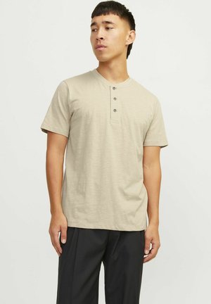 Camiseta henley de manga corta beige con tapeta de tres botones. Tela suave y texturizada con un corte relajado. Llevada con pantalones oscuros.